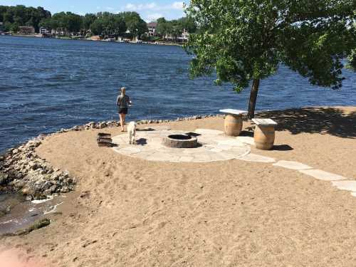 A person walks a dog along a sandy shore by a lake, with a fire pit and trees in the background.