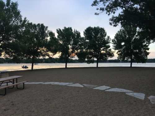 A serene lakeside view with trees lining the shore and a picnic table in the foreground, under a colorful sunset sky.