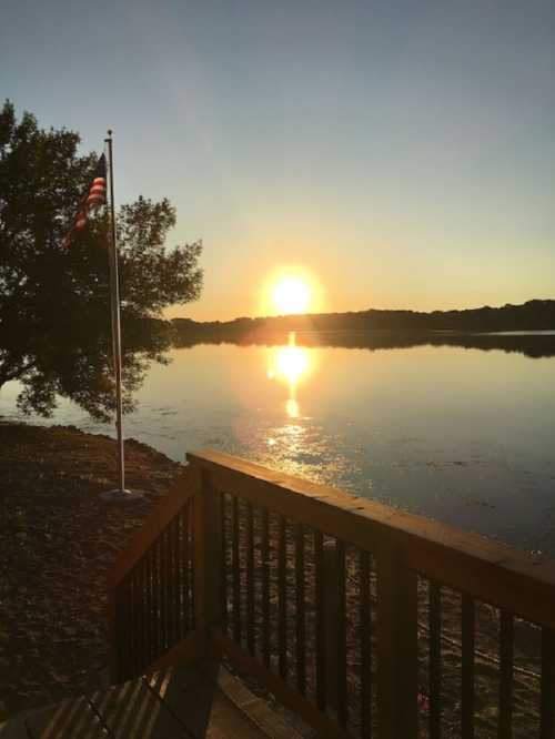 A serene sunset over a calm lake, with an American flag waving nearby and trees silhouetted against the sky.