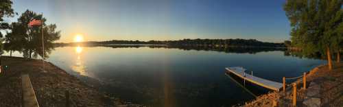 Panoramic view of a calm lake at sunrise, with a flag and dock on the shore, surrounded by trees.