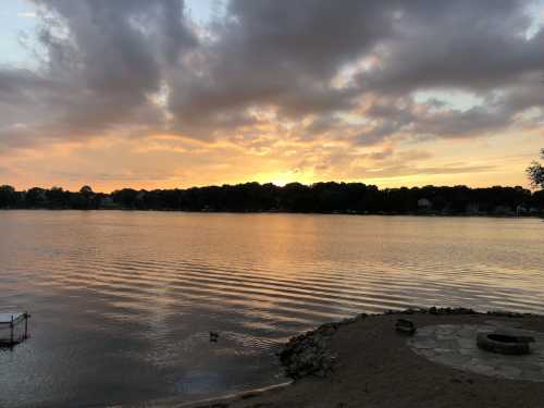 Sunset over a calm lake, with rippling water and a tree-lined shore under a cloudy sky.