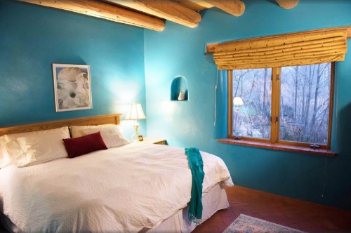 Cozy bedroom with a blue wall, a large bed, a lamp, and a window overlooking a natural landscape.