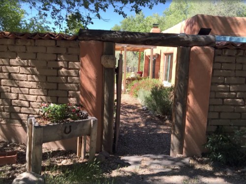 A rustic entrance with a wooden gate leads to a garden path surrounded by greenery and adobe-style buildings.