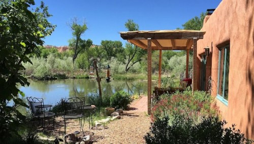 A serene outdoor scene featuring a pond, greenery, and a cozy patio with chairs under a wooden pergola.