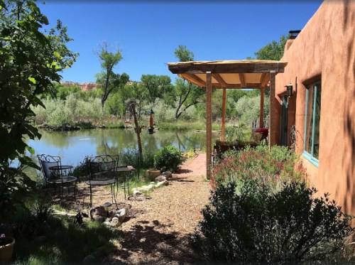 A serene outdoor scene featuring a pond, greenery, and a cozy patio with chairs under a wooden pergola.