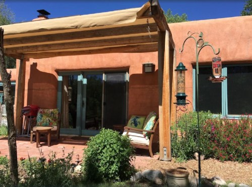 A cozy patio with a wooden awning, colorful cushions, and a bird feeder, surrounded by greenery and flowers.