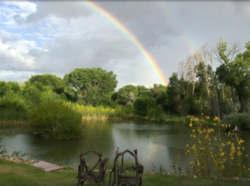 A serene pond surrounded by greenery, with a vibrant double rainbow arching across a cloudy sky. Two chairs in the foreground.