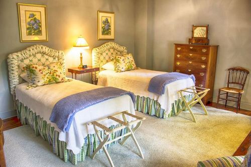 A cozy bedroom with two twin beds, a dresser, and a lamp, featuring floral decor and a soft rug.