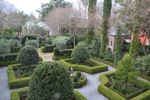 A lush garden with neatly trimmed hedges, topiary, and pathways, surrounded by trees and buildings in the background.