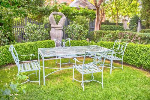 A glass-top dining table surrounded by four metal chairs in a lush garden with greenery and a stone sculpture.
