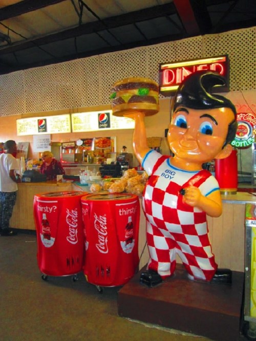 A large statue of a boy in checkered overalls holding a burger, next to a diner counter with Coca-Cola displays.