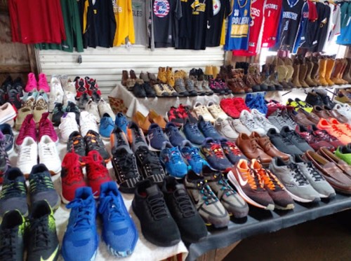 A variety of shoes displayed on tables, including sneakers, boots, and casual footwear, with clothing in the background.
