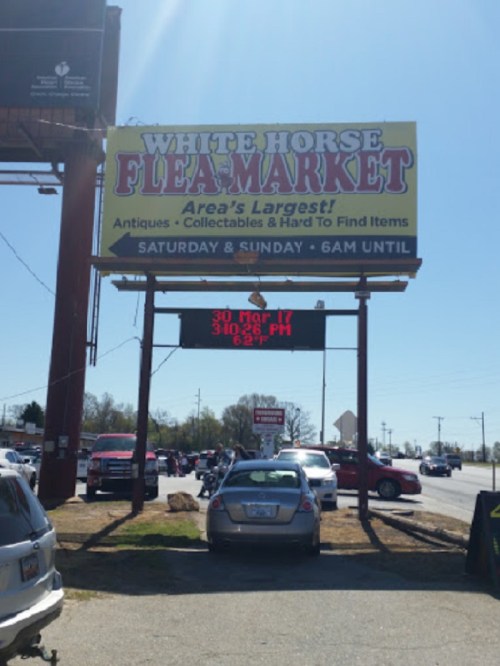 Sign for White Horse Flea Market, advertising hours and date, with cars parked nearby on a sunny day.