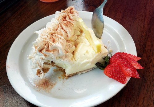 A slice of creamy dessert topped with whipped cream and a strawberry, served on a white plate with a spoon.