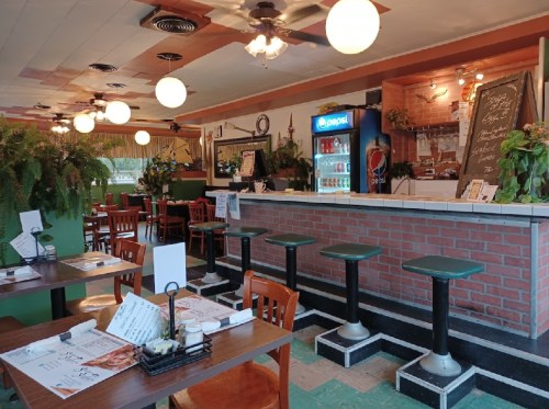 Cozy restaurant interior with wooden tables, green walls, a bar area, and hanging plants. Bright lighting and a soda fridge visible.
