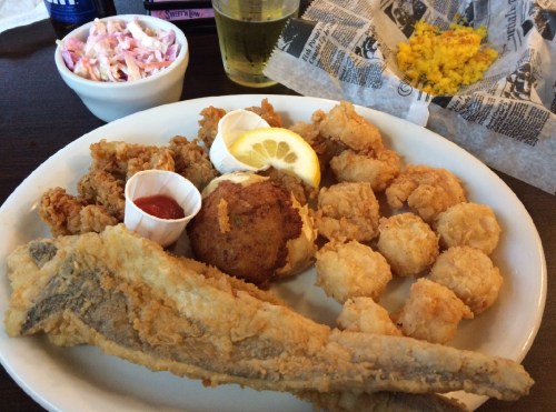 A plate of fried fish, shrimp, hushpuppies, and coleslaw, with a lemon wedge and dipping sauce on the side.