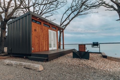 A modern tiny house with wooden and metal exterior, set by a lake, surrounded by trees and a grill nearby.