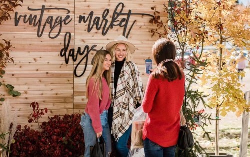Two women pose for a photo in front of a "Vintage Market Days" sign, surrounded by autumn leaves and decor.