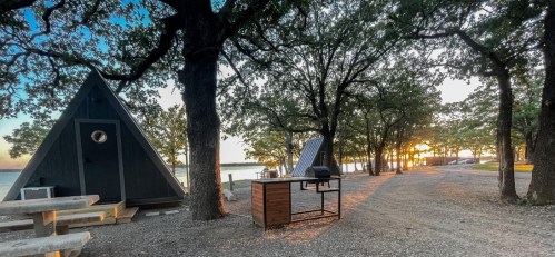 A serene lakeside scene featuring A-frame cabins, picnic tables, and a sunset through trees along a gravel path.