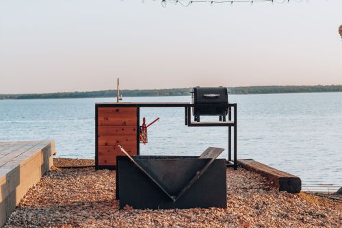 A modern outdoor grilling station by a lake, featuring a grill, storage drawers, and a fire pit on a gravel surface.