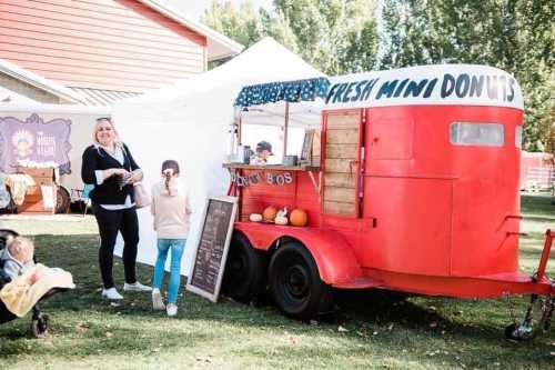 A woman stands by a red donut trailer with a sign reading "Fresh Mini Donuts," surrounded by a festive outdoor setting.