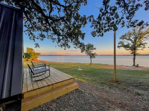 A serene lakeside view with a wooden deck, two chairs, and trees under a colorful sunset sky.
