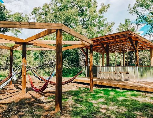 A wooden structure with swings and colorful hammocks under trees, featuring a shaded area with a metal roof.