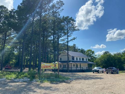 A building surrounded by trees with a "Now Open" sign in front and vehicles parked nearby on a gravel lot.