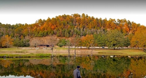 A person stands by a calm lake, reflecting autumn trees and a hillside in warm colors.