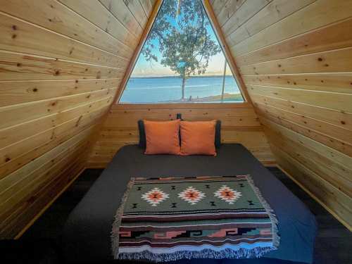 Cozy triangular cabin interior with a bed, orange pillows, and a view of the lake through large windows.