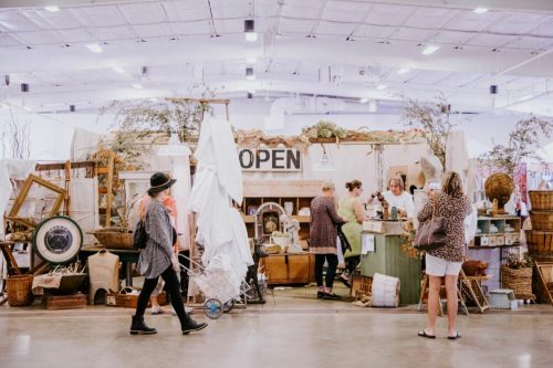 A bustling market scene with shoppers browsing vintage items at a cozy, open booth filled with decor and plants.
