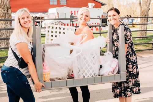 Three women smiling while carrying a large cart filled with bags and items outdoors.