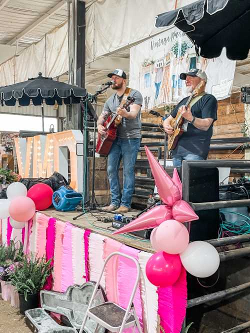 Two musicians perform on stage with guitars, surrounded by colorful decorations and a festive atmosphere.