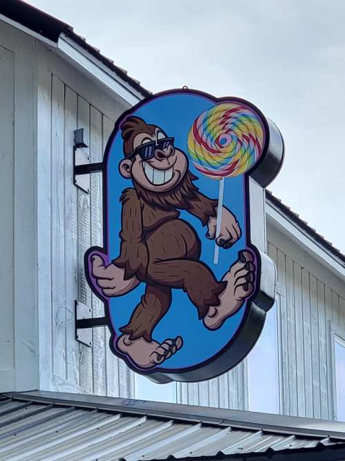 A cartoonish Bigfoot character wearing sunglasses, holding a colorful lollipop, on a blue background.