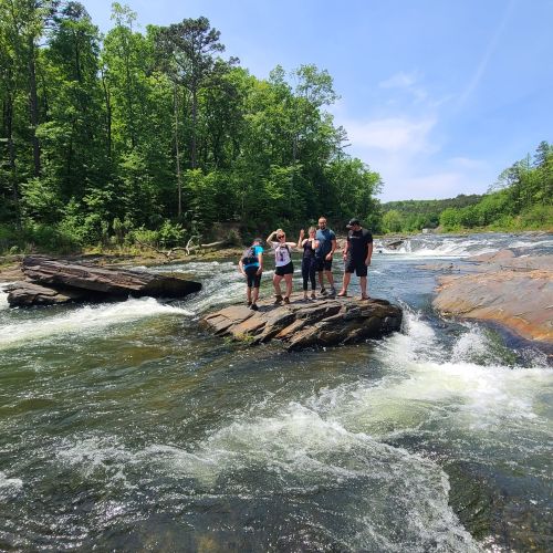 A group of people standing on rocks by a river, surrounded by trees and a clear blue sky.
