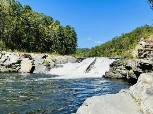 A waterfall cascades over rocky ledges, surrounded by lush green trees under a clear blue sky.
