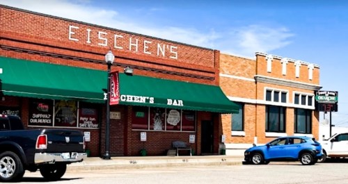 Exterior of Eischen's Bar, a brick building with green awnings, featuring a parked blue car and a sunny sky.
