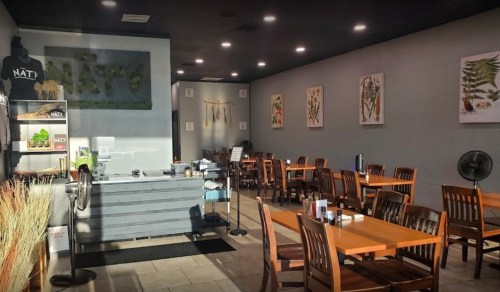 Interior of a restaurant featuring wooden tables, chairs, and wall art, with a service counter in the foreground.