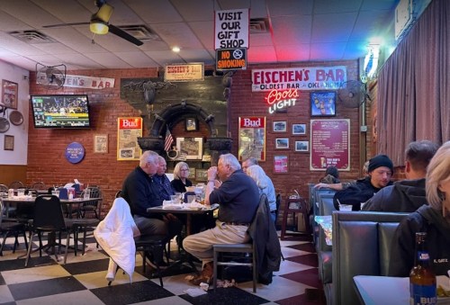 A cozy bar scene with patrons dining and chatting, featuring vintage decor and a TV showing a sports game.