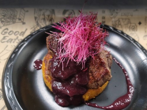 A plated dish featuring a piece of meat topped with pink microgreens and a dark berry sauce on a corn base.