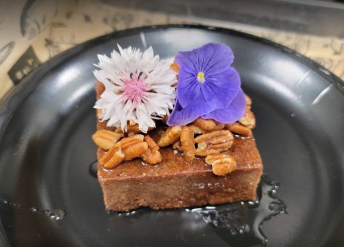 A square dessert topped with nuts and edible flowers on a black plate, drizzled with syrup.