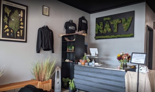 Interior of a shop featuring a moss-covered sign, plants, merchandise, and a reception desk with flowers.