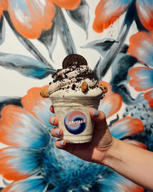 A hand holds a cup of cookies and cream ice cream topped with an Oreo, against a colorful floral mural background.