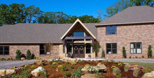 A modern brick house with a large front porch, surrounded by landscaped gardens and trees under a clear blue sky.
