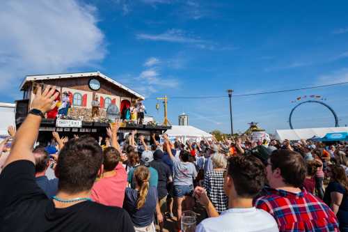 A lively crowd raises their hands at an outdoor event with a stage, performers, and a Ferris wheel in the background.