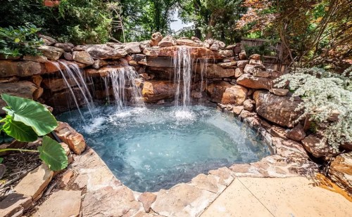 A serene outdoor hot tub surrounded by natural stone, with cascading waterfalls and lush greenery.