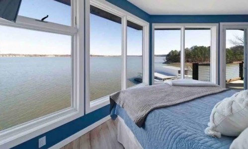 A serene bedroom with large windows overlooking a calm lake and distant trees, featuring a cozy bed and blue walls.