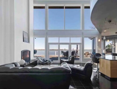 Modern living room with large windows overlooking a waterfront, featuring black leather furniture and a sleek design.