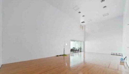 A spacious, empty indoor court with wooden flooring and white walls, featuring a glass-walled area in the background.