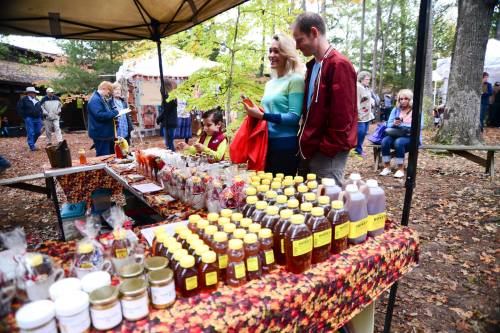A couple admires honey jars at a fall market, surrounded by colorful decorations and other vendors in the background.